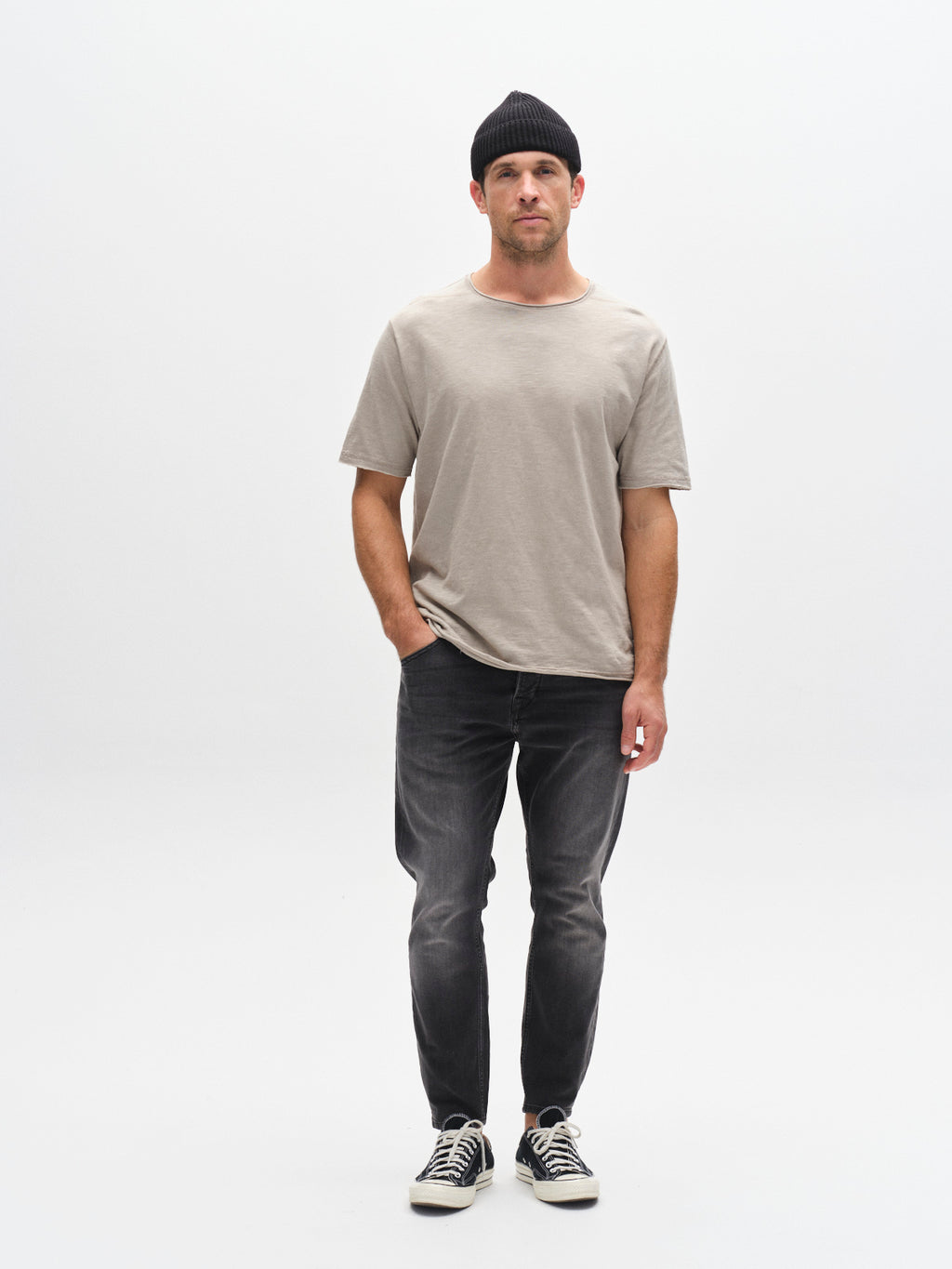 A man stands against a white background, wearing a black beanie, loose beige t-shirt, GABBA GABalex Thor Cropped Jeans in denim wash, and black-and-white sneakers. One hand in his pocket, he has a neutral expression.