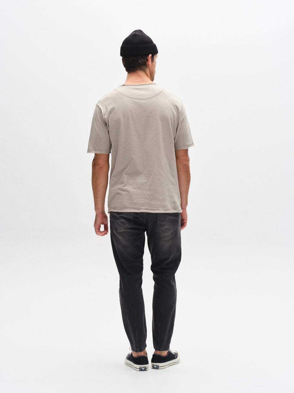 A man faces away from the camera in a beige T-shirt, black beanie, slip-on shoes, and GABBA's GABalex Thor Cropped Jeans in Denim wash. The background is plain white.