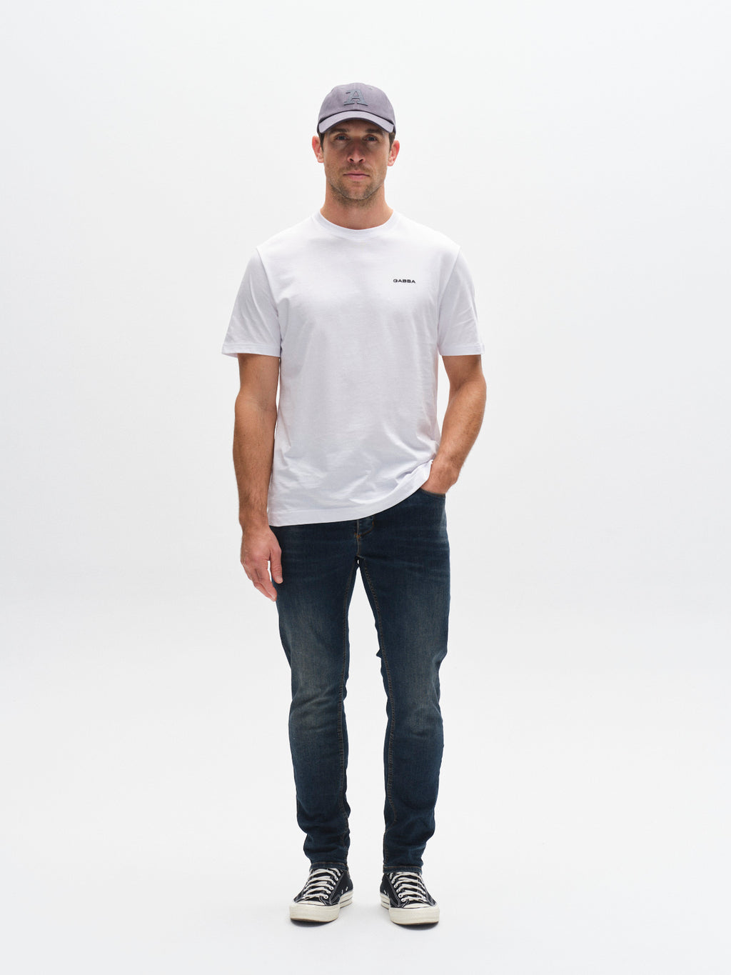 A man in a light gray cap, plain white t-shirt, GABBA GABrey K3606 Mid Blue Jeans in mid blue denim, and black-and-white sneakers stands against a white background with one hand in his pocket, highlighting a casual style.