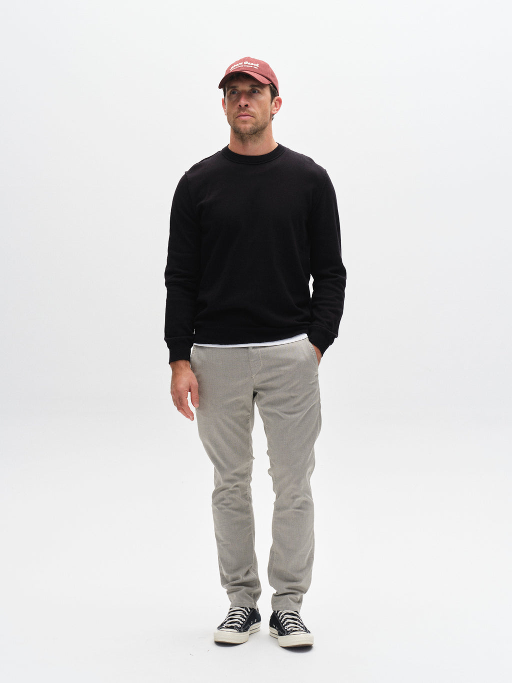 A man stands against a white background wearing a red baseball cap, black sweater, black-and-white sneakers, and Gabba’s GABpaul K3280 Dale Chino - Birch with French pockets and double-paspel back pockets. Left hand in pocket, neutral expression.