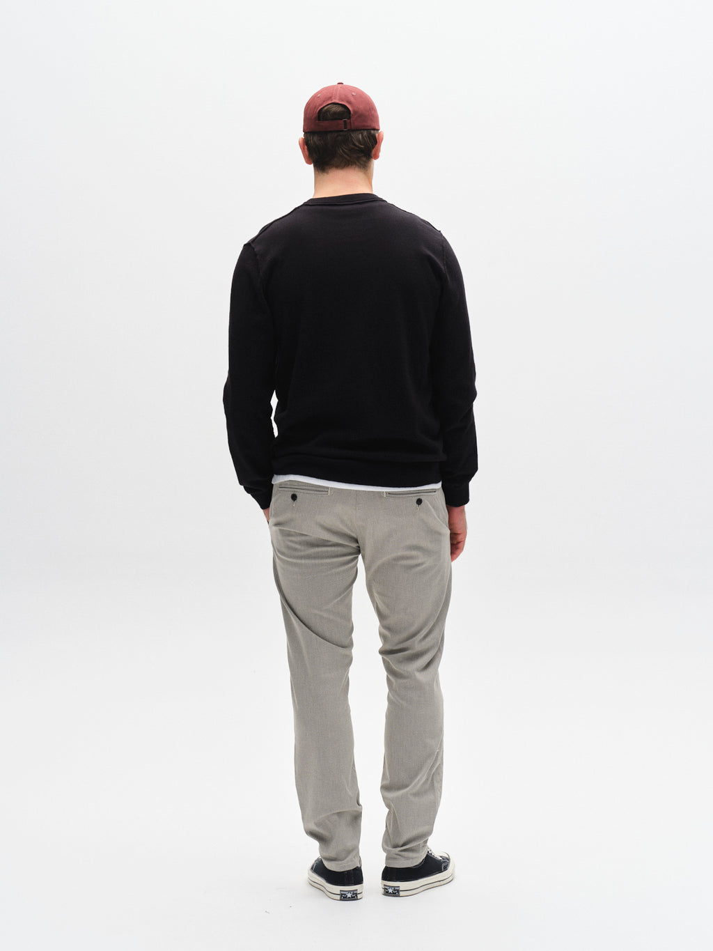 A man faces away from the camera, wearing a red cap, black sweatshirt, Gabba GABpaul K3280 Dale Chino - Birch pants with French pockets, and dark slip-on shoes against a plain white background.