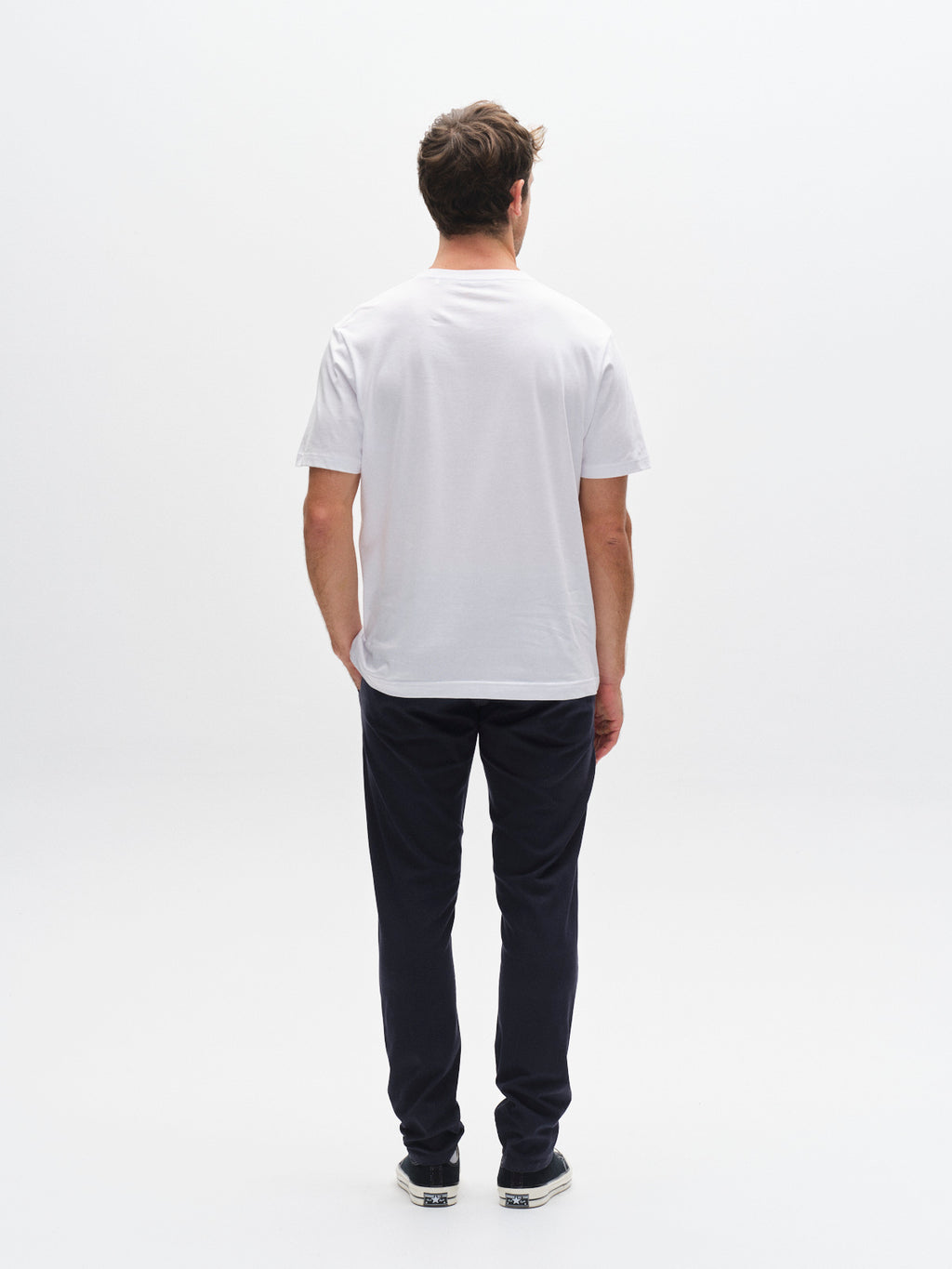 A man stands facing away from the camera against a white background, wearing a white short-sleeve t-shirt, GABBA GABpaul K3280 Dale Chino - Navy pants with French pockets, and sneakers.