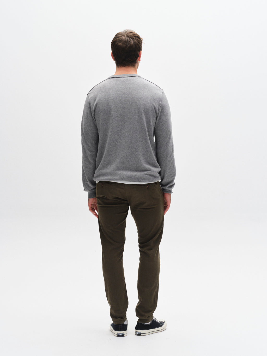 A man faces away from the camera against a white background, wearing a gray long-sleeve shirt and GABBA GABpaul K3280 Dale Chino - Army pants with French pockets, paired with black sneakers featuring white soles.