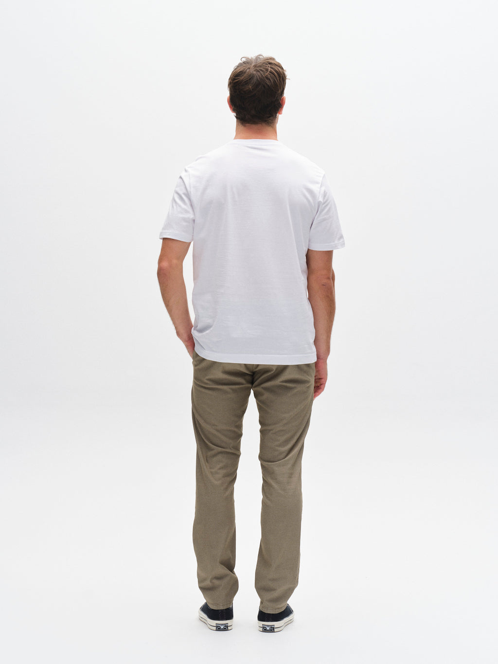 A man stands facing away from the camera in a plain white T-shirt, black sneakers, and Gabba GABpaul K3280 Dale Chino in Olive Green, with French pockets, against a white background.