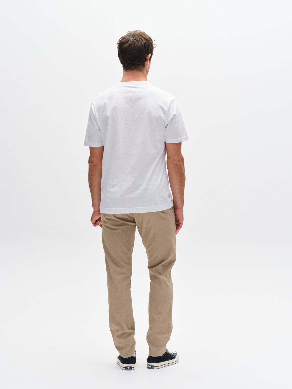 A man stands facing away from the camera, wearing a plain white T-shirt, Gabba GABpaul K3280 Dale Chino in sand, and black sneakers against a white background.