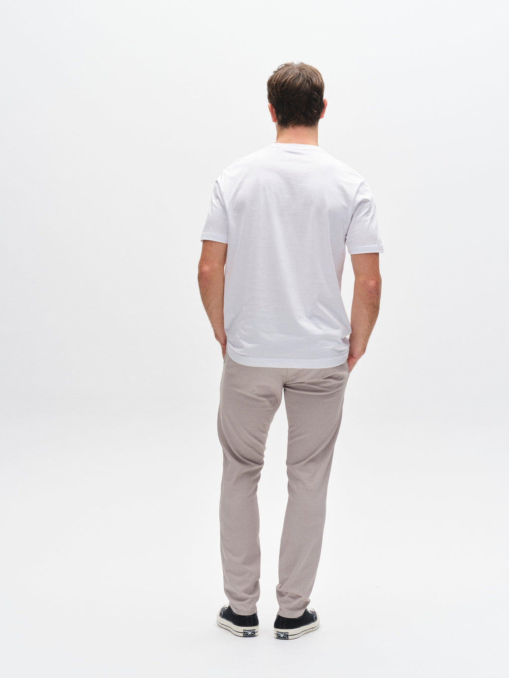 A man stands facing away from the camera, wearing a plain white T-shirt, Gabba GABpaul K3280 Dale Chino in Grey Paloma with French and double-paspel back pockets, paired with black sneakers, against a white background.
