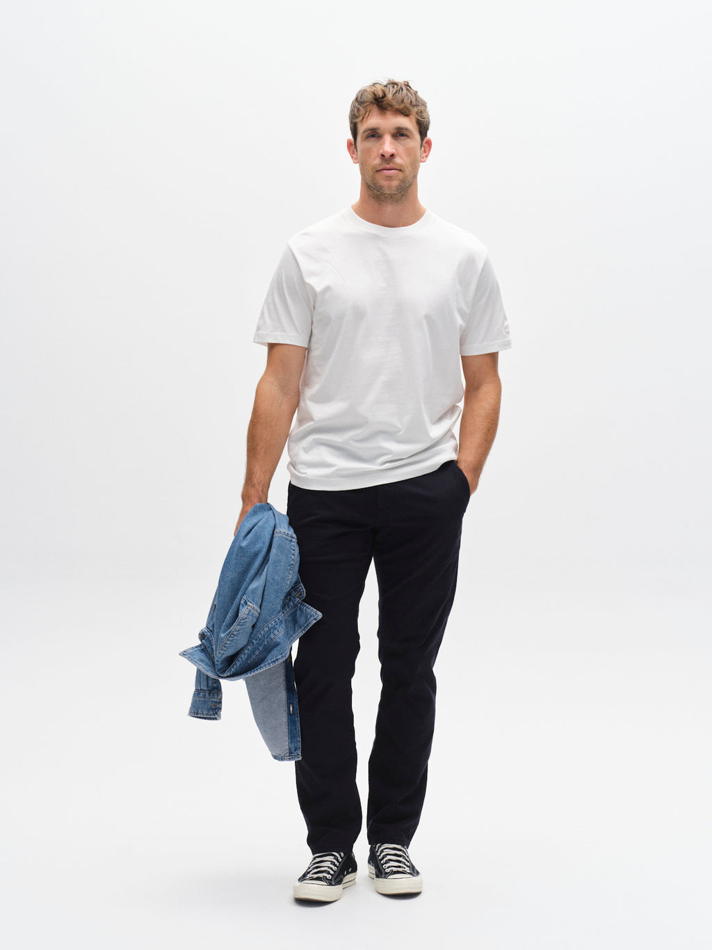 A man stands against a white background, wearing a white t-shirt, Gabba GABpaul K3280 Dale Chino in Blue Black with French pockets, black sneakers, and holding a blue denim jacket in his left hand for a classic style.