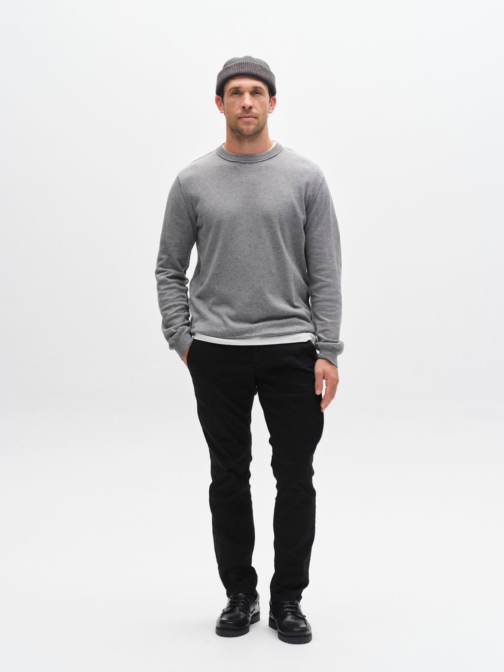 A man stands facing forward against a white background, wearing a gray sweater, black GABBA GABpaul K3280 Dale Chino pants with French pockets, black shoes, and a gray beanie. His left hand is in his pocket and his expression is neutral.