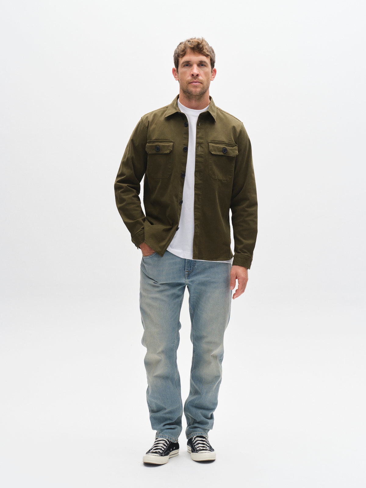 A man faces forward against a plain white background, wearing the GABBA GABtopper LS Shirt - Army with chest pockets and a shirt collar over a white t-shirt, paired with light blue jeans and black-and-white sneakers.