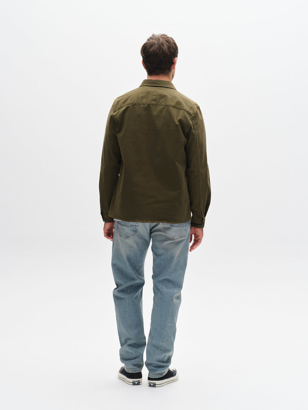 A man faces away from the camera against a white background, wearing the GABBA GABtopper LS Shirt - Army with chest pockets, paired with light blue jeans and black-and-white sneakers.