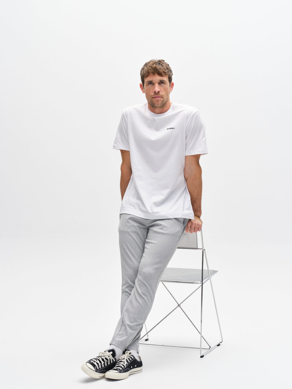 A man in a white t-shirt, Gabba Pisa Domo Pants in Monument, and black sneakers leans against a metal chair in a minimal, white studio setting.