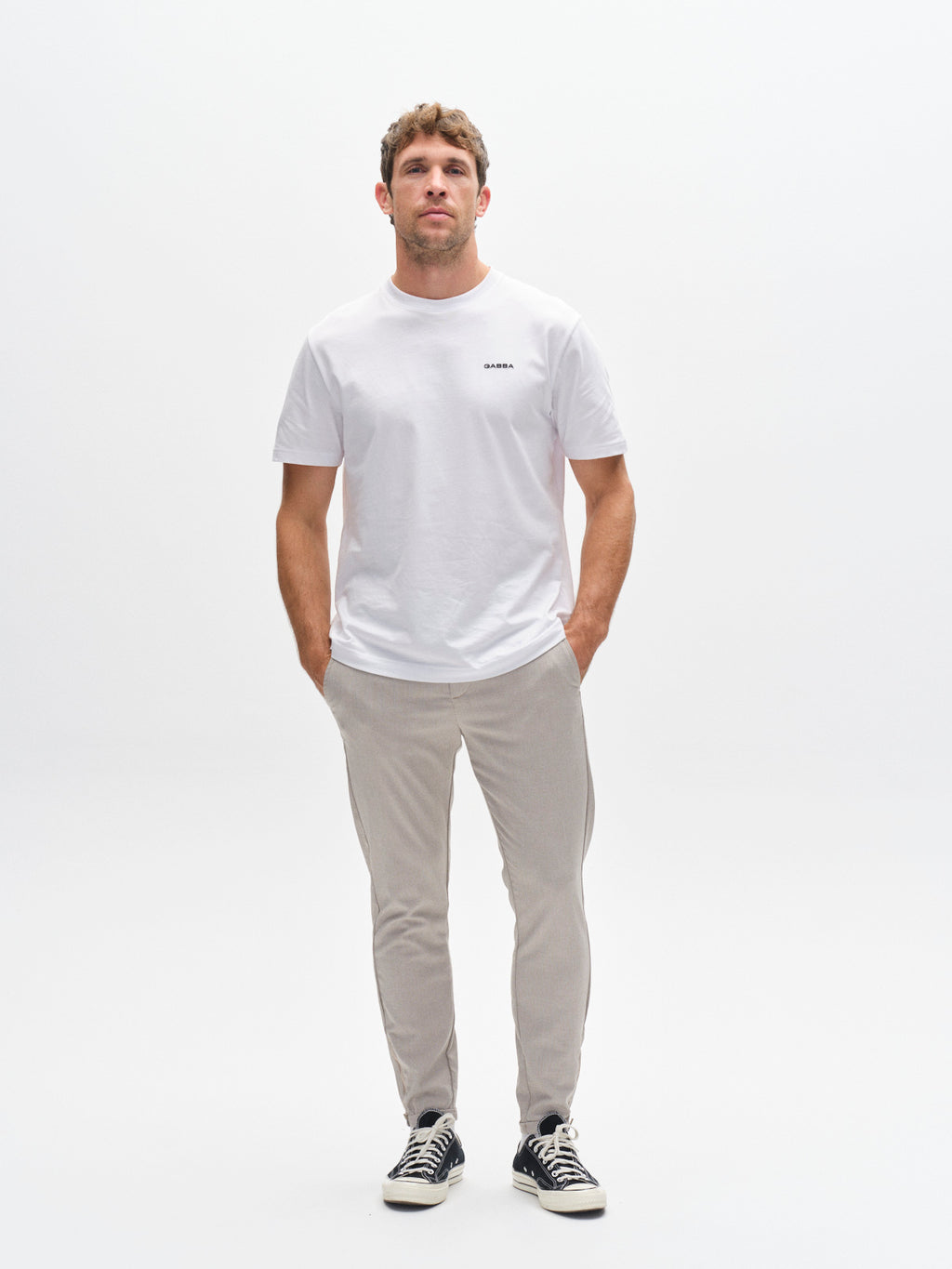 A man stands facing forward with his hands in his pockets, wearing a test-print white T-shirt, Gabba Pisa Domo Pants in Coriander, and black sneakers against a plain white background.