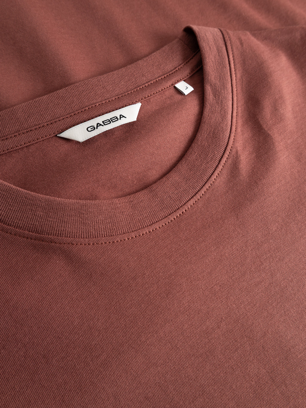 Close-up of a folded, brick red organic cotton GABdune Logo GOTS SS T-Shirt from Gabba, featuring a white label reading Gabba and a small size tag marked L near the collar.