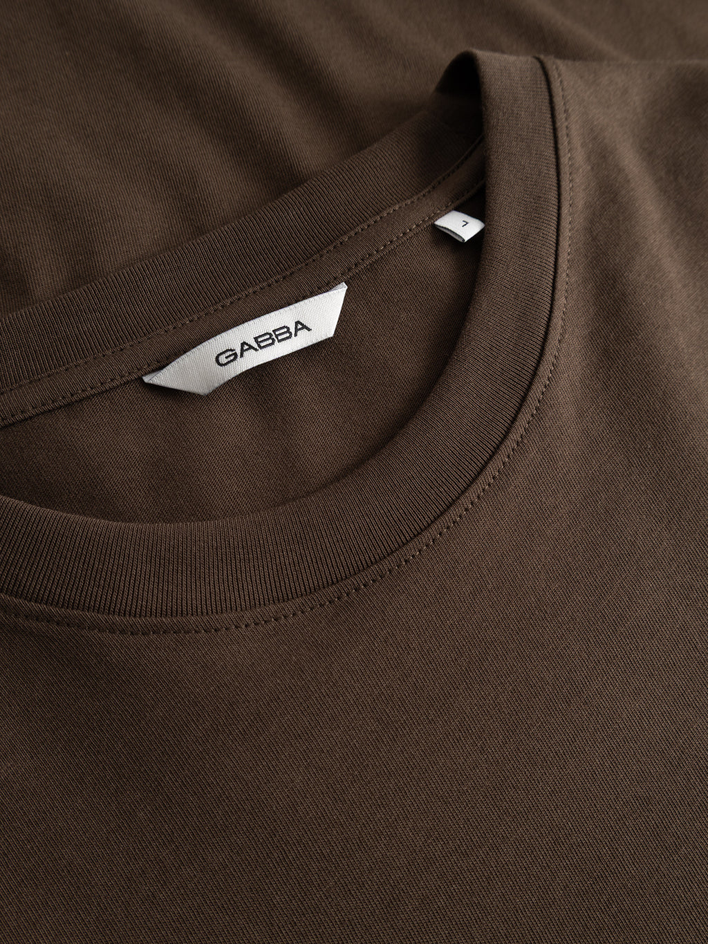 Close-up of the GABdune Logo GOTS SS T-Shirt in Brown Slate, a classic fit piece by Gabba. The shirt is neatly folded to showcase the neckline and fabric texture, with a white inner label bearing the brand name Gabba in black text.