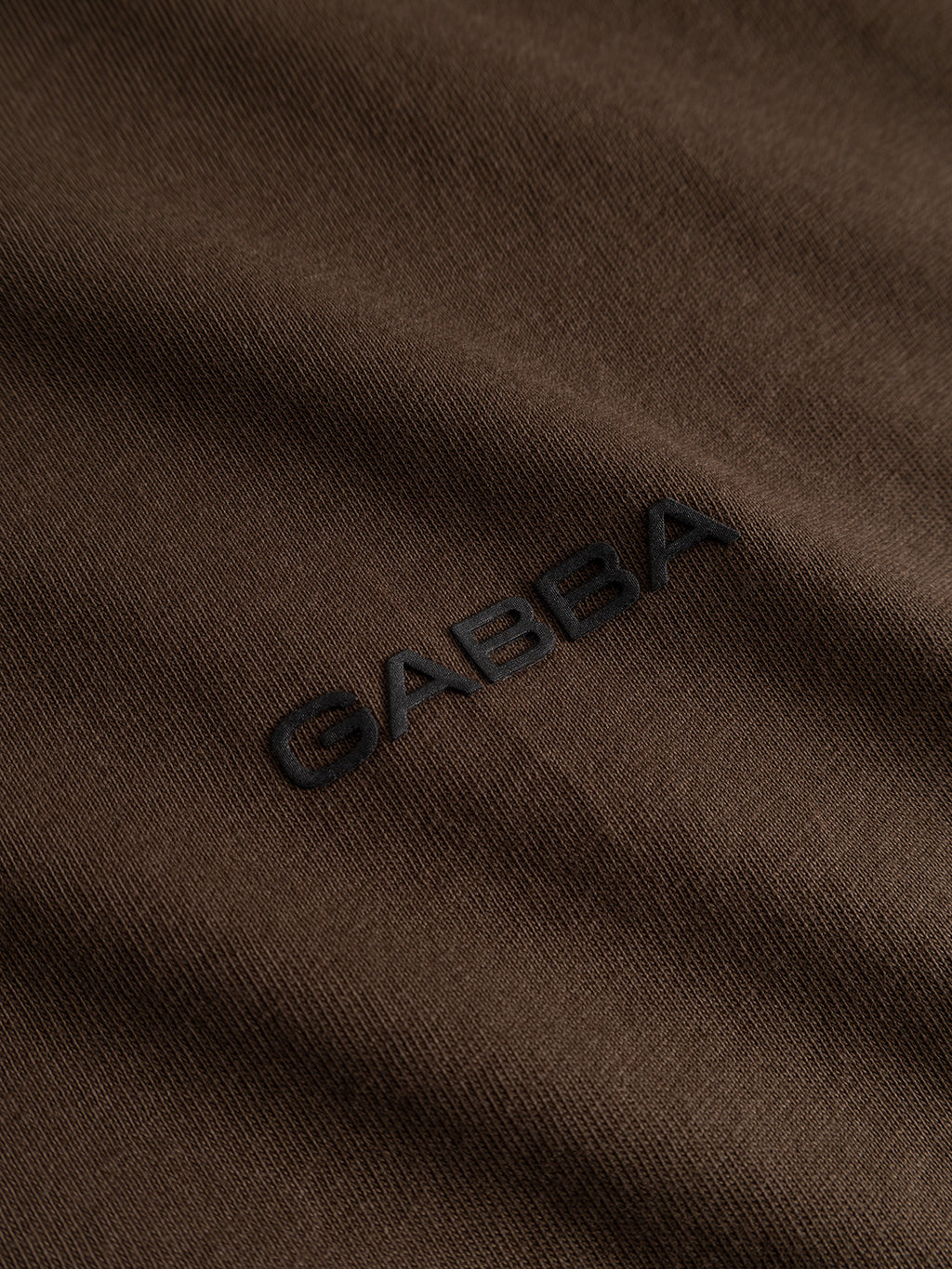 Close-up of brown slate fabric with the Gabba logo “GABBA” stitched in raised black letters. The smooth, finely woven texture highlights the premium quality of the classic fit GABdune Logo GOTS SS T-Shirt, crafted from GOTS-certified organic cotton by Gabba.