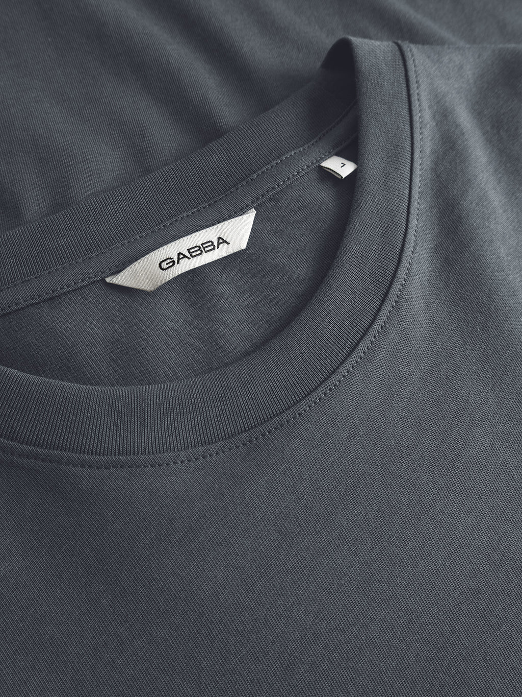 Close-up of a Turbulence Blue crew neck classic fit t-shirt with a visible white fabric label that reads Gabba. Expertly crafted from organic cotton, this GOTS-certified GABdune Logo SS T-Shirt is neatly folded, highlighting the neckline and part of the chest area.