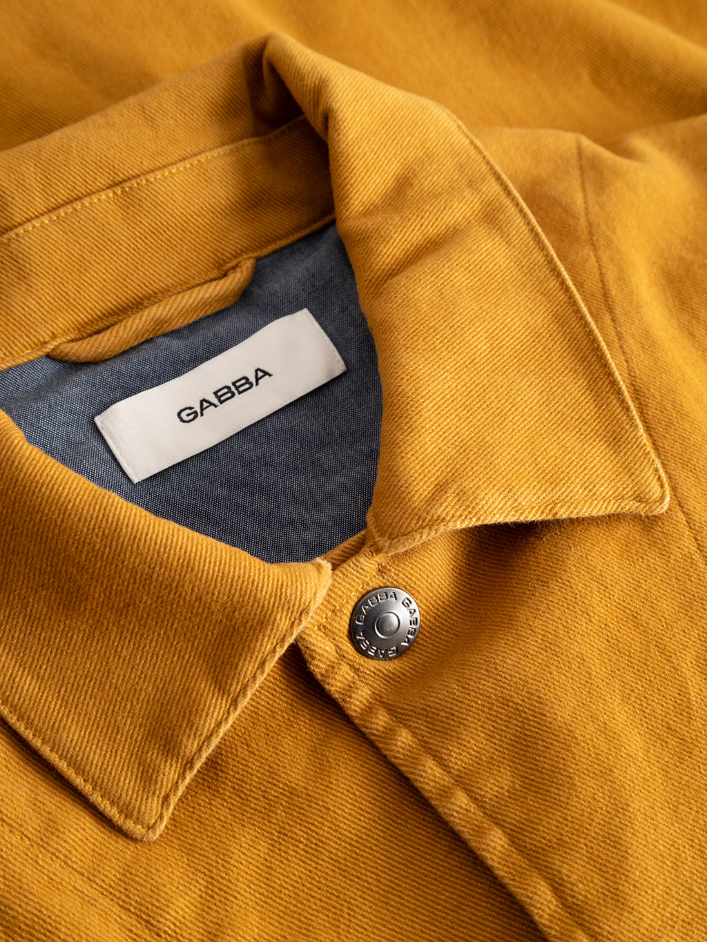 Close-up of the GABbeta Twill jacket by Gabba in mustard yellow, crafted from garment-dyed stretch twill. This regular fit design features utility pockets, a silver button, and a white label inside the collar that reads “GABBA.” The inner lining is gray.
