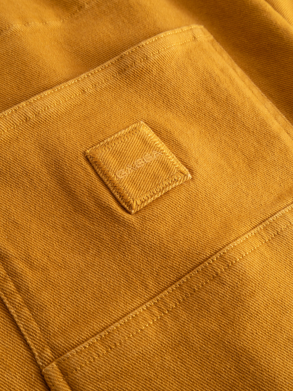A close-up of a GABbeta Twill - Mustard Yellow utility pocket by Gabba, showcasing the garment-dyed stretch twill fabric with a small stitched square patch in the center. The texture and detailed stitching of the fabric are clearly visible.
