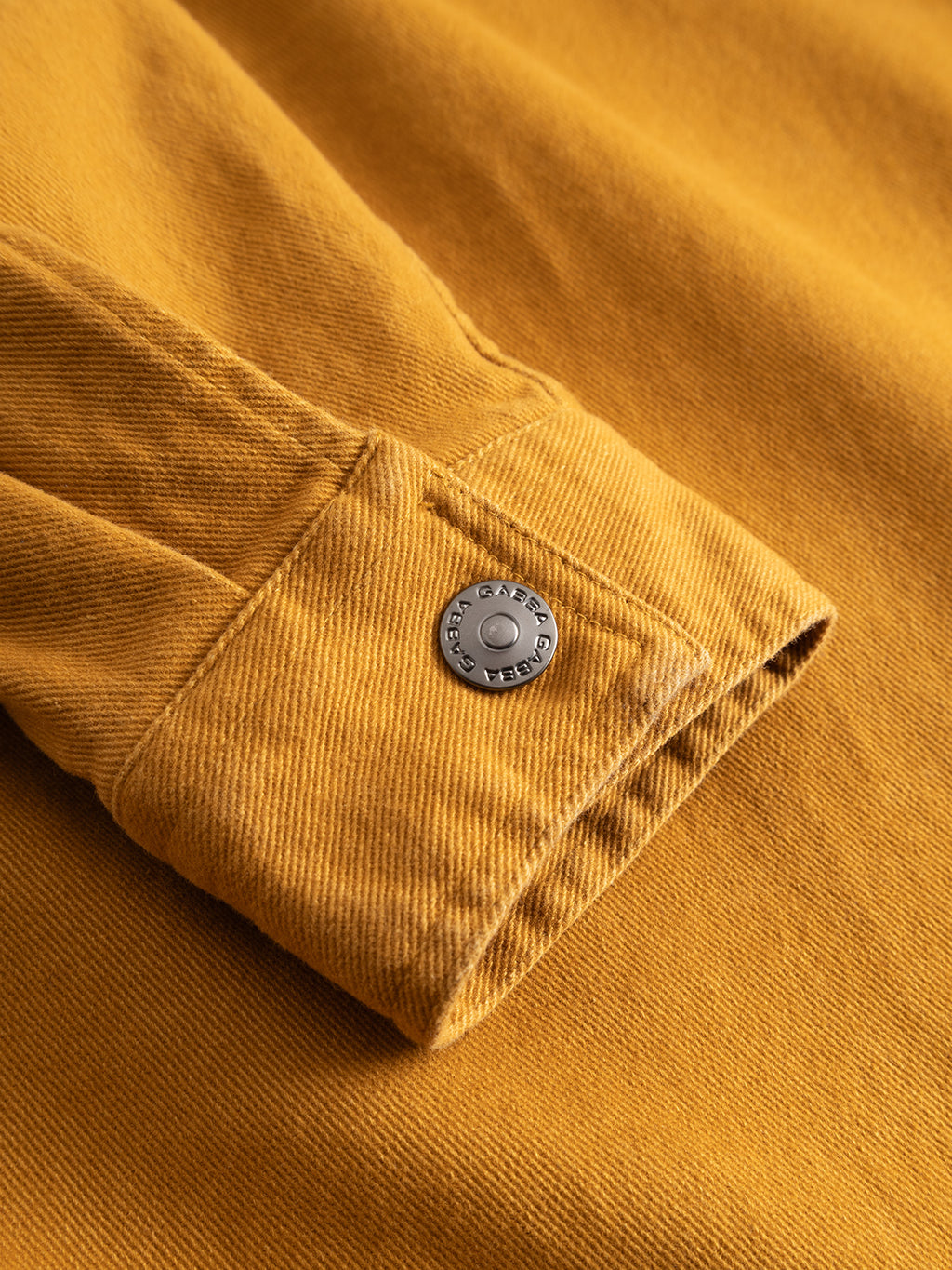A close-up of a mustard yellow GABbeta Twill sleeve by Gabba, featuring a buttoned cuff with a metal button engraved “GARMENT.” The textured fabric is garment-dyed stretch twill, and thoughtful utility pocket-inspired details add practicality.