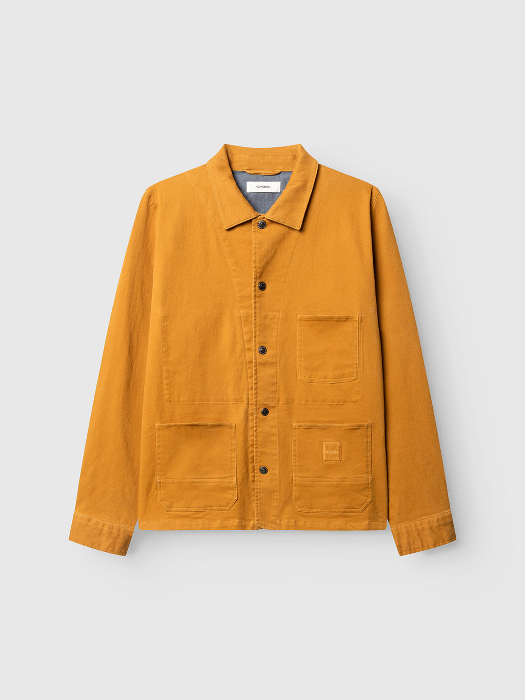 The Gabba GABbeta Twill in mustard yellow is a regular fit work jacket made from garment-dyed stretch twill, designed with a collar, front button closure, and three utility pockets, and is shown on a plain light gray background.