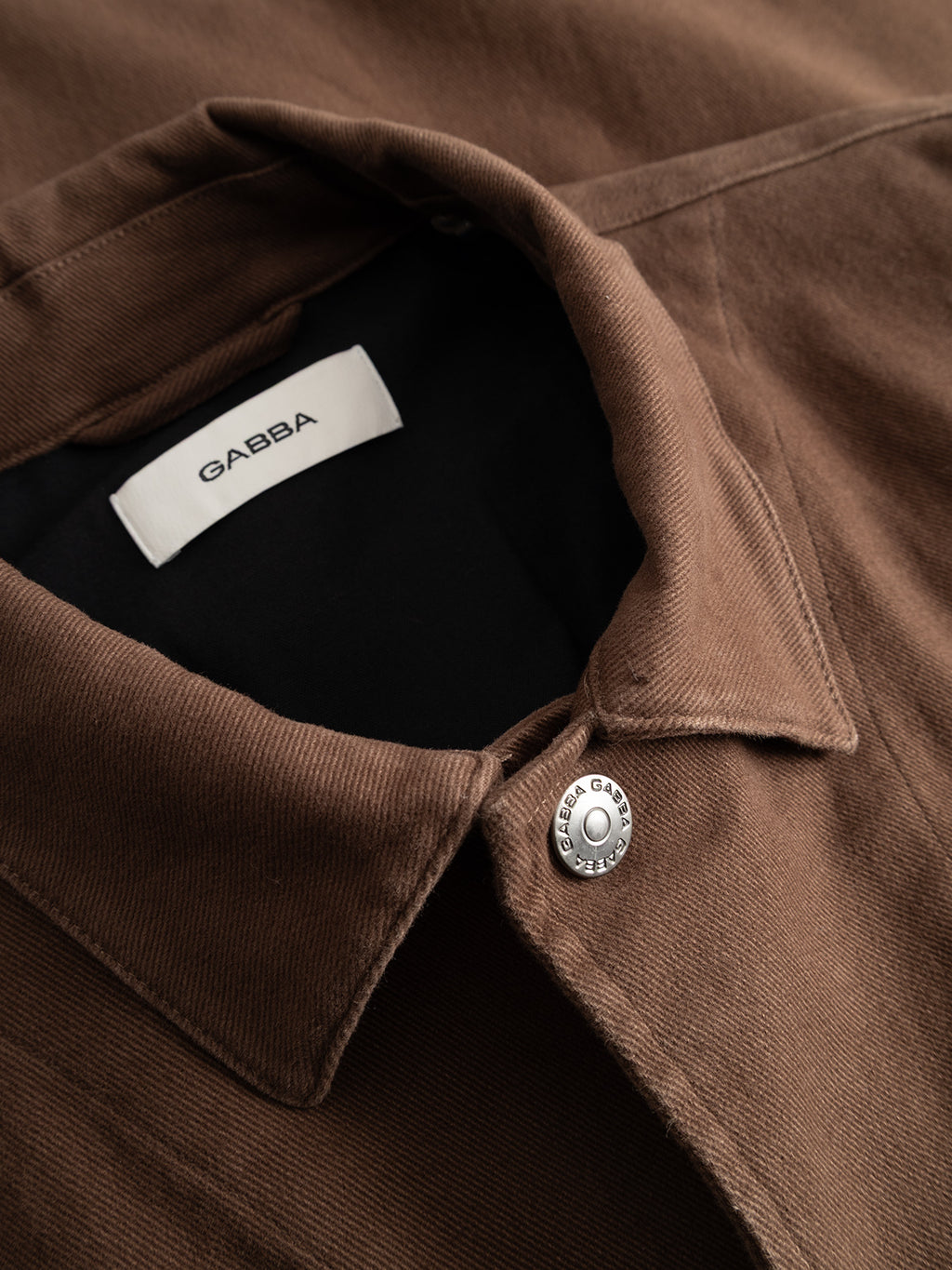 Close-up of the Gabba GABbeta Twill - Brown Slate jacket, featuring a workwear-inspired silhouette in brown twill fabric, a silver button engraved with “GABBA,” and a white inner label bearing the Gabba name.