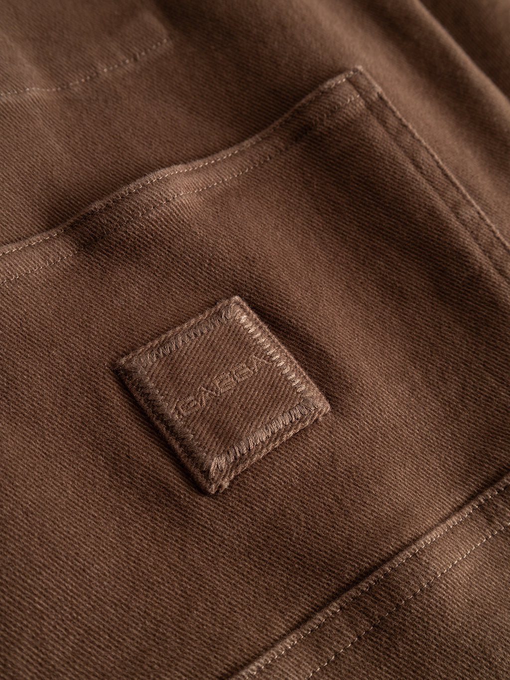 Close-up of a brown fabric pocket with a small stitched square patch above it. Made from Gabba’s GABbeta Twill in Brown Slate, this garment-dyed stretch twill features a textured, durable finish and visible stitching details that emphasize its workwear-inspired silhouette.