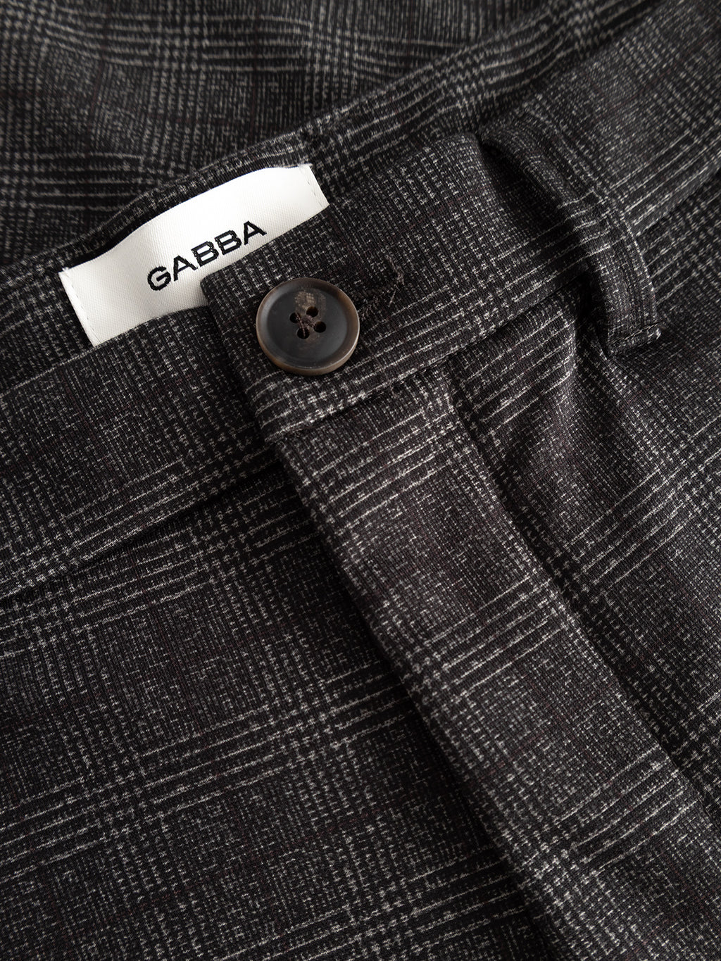 Close-up of GABpaul Nygo - Grey Check pants by Gabba, featuring a black and white plaid pattern with a brown button and a white GABBA label inside the waistband. These slim chino-fit pants are crafted from comfort stretch fabric, making them ideal for everyday wear.