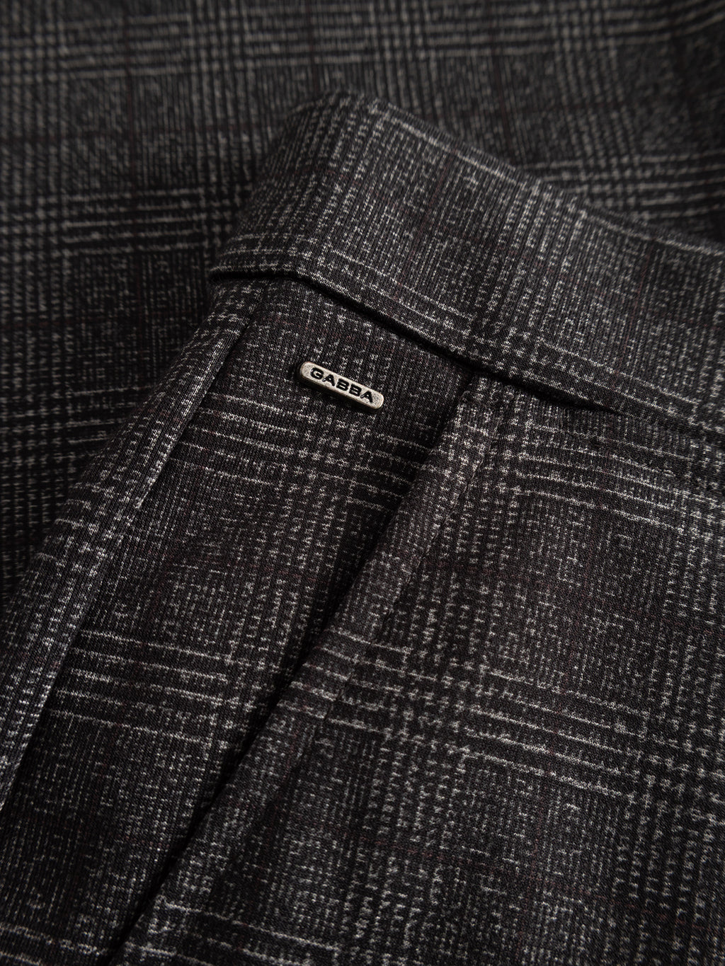 Close-up of the grey check fabric from GABpaul Nygo by Gabba, featuring a small metallic label embossed with “DABBA” near the seam. The photo highlights the detailed textile texture and pattern—crafted from comfort stretch fabric, making it perfect for everyday wear.