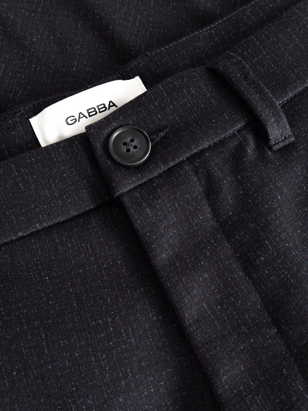Close-up of a pair of dark textured pants with a black button, belt loop, and a visible white label that reads Gabba. The GABpaul Nygo - Night Sky chinos by Gabba are crafted from comfort stretch fabric, offering versatile style and ease for everyday wear.