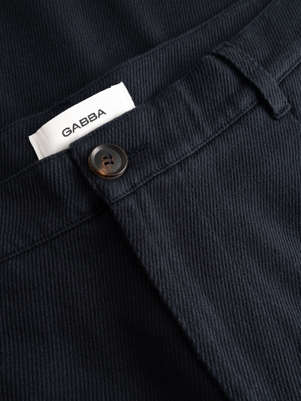 Close-up of the waistband of Gabba GABJoe-Pas Vill trousers in Blue Black, featuring a black button and a white GABBA label. The drapey cotton twill fabric texture and utility-inspired stitching details are clearly visible.