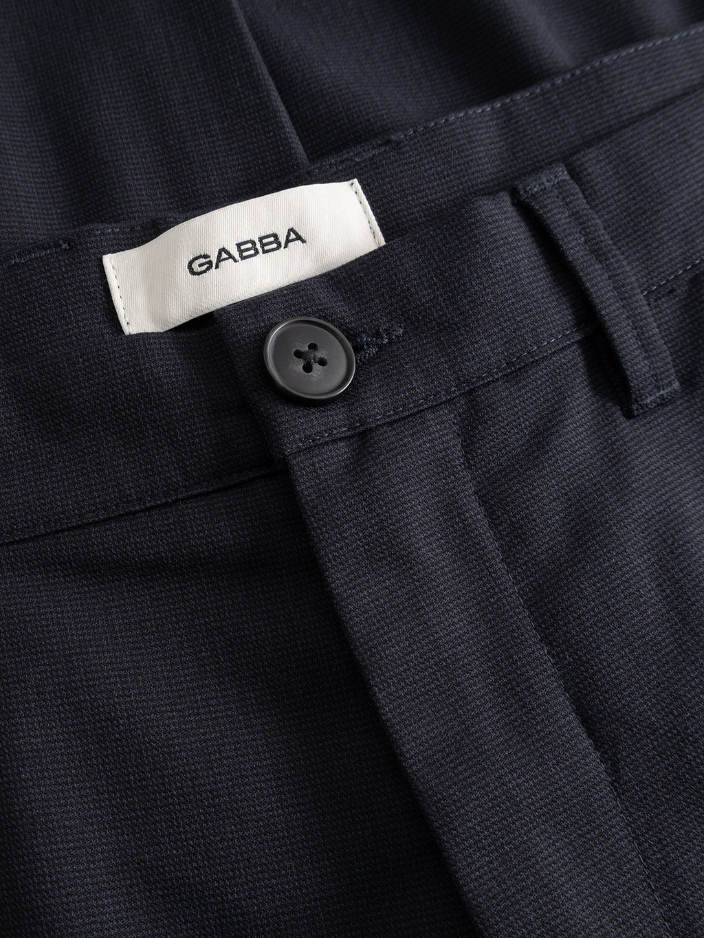Close-up of the GABpaul Deme Pant in dark navy by Gabba, crafted from a micro-pattern woven fabric and featuring a black button, belt loops, and a white label with “GABBA” sewn inside the waistband. This slim chino fit is perfect for everyday wear.
