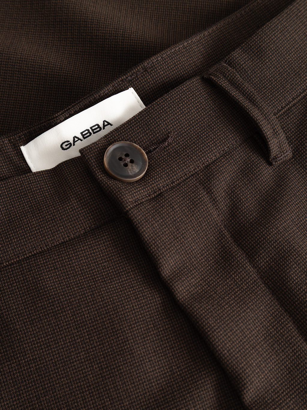 Close-up of the GABpaul Deme Pant in Brown Slate by Gabba, featuring a brown micro-pattern woven fabric, a four-hole brown button, and a white waistband label reading “Gabba,” highlighting the pants’ versatile chino style.