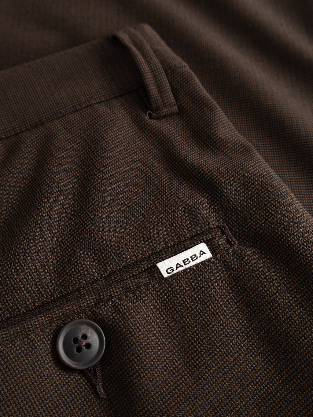 Close-up of the Gabba GABpaul Deme Pant in Brown Slate, showcasing a buttoned back pocket with a small white Gabba label above it. The fabric displays a micro-pattern woven texture, adding subtle detail to these versatile chino pants.