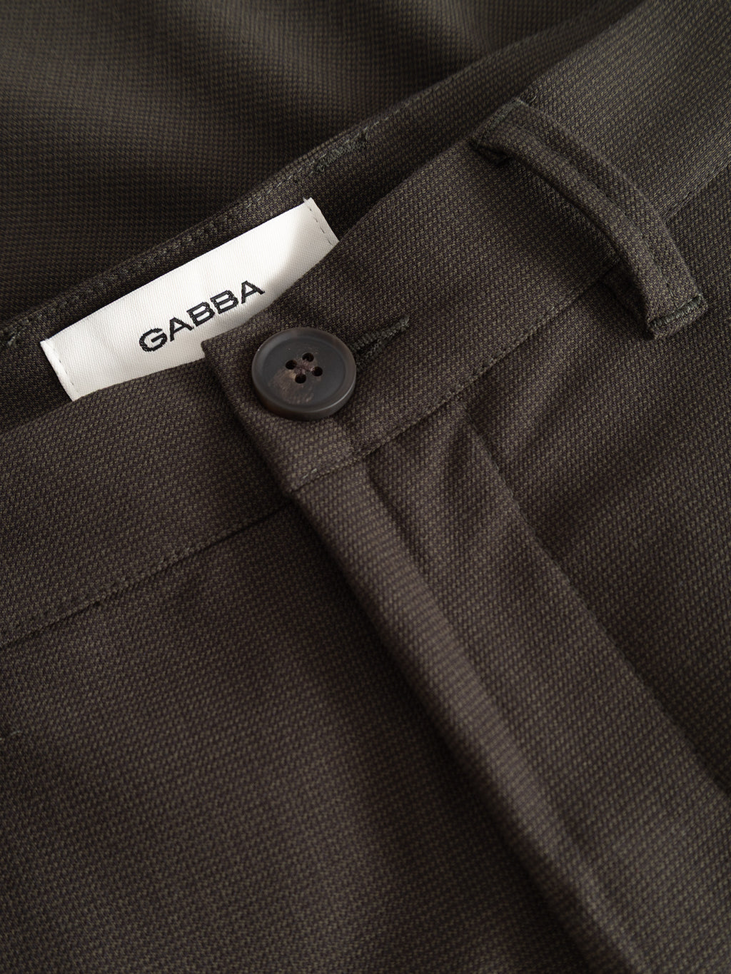 Close-up of the GABpaul Deme Pant in Forest Night, featuring dark textured fabric woven with a subtle micro-pattern and a black button securing the waistband. Inside the waistband, a white label showcases the Gabba brand name in bold black letters.