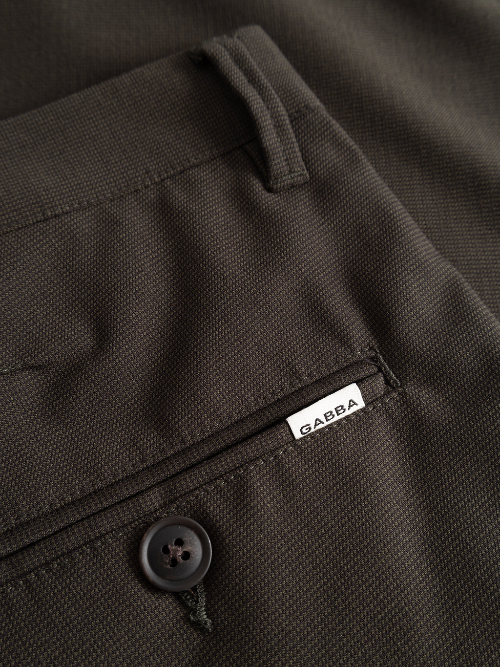 Close-up of the GABpaul Deme Pant in Forest Night by Gabba, showcasing the dark brown micro-pattern woven fabric, a back pocket with a black button, and a small white Gabba label. These versatile pants feature a modern slim chino fit.