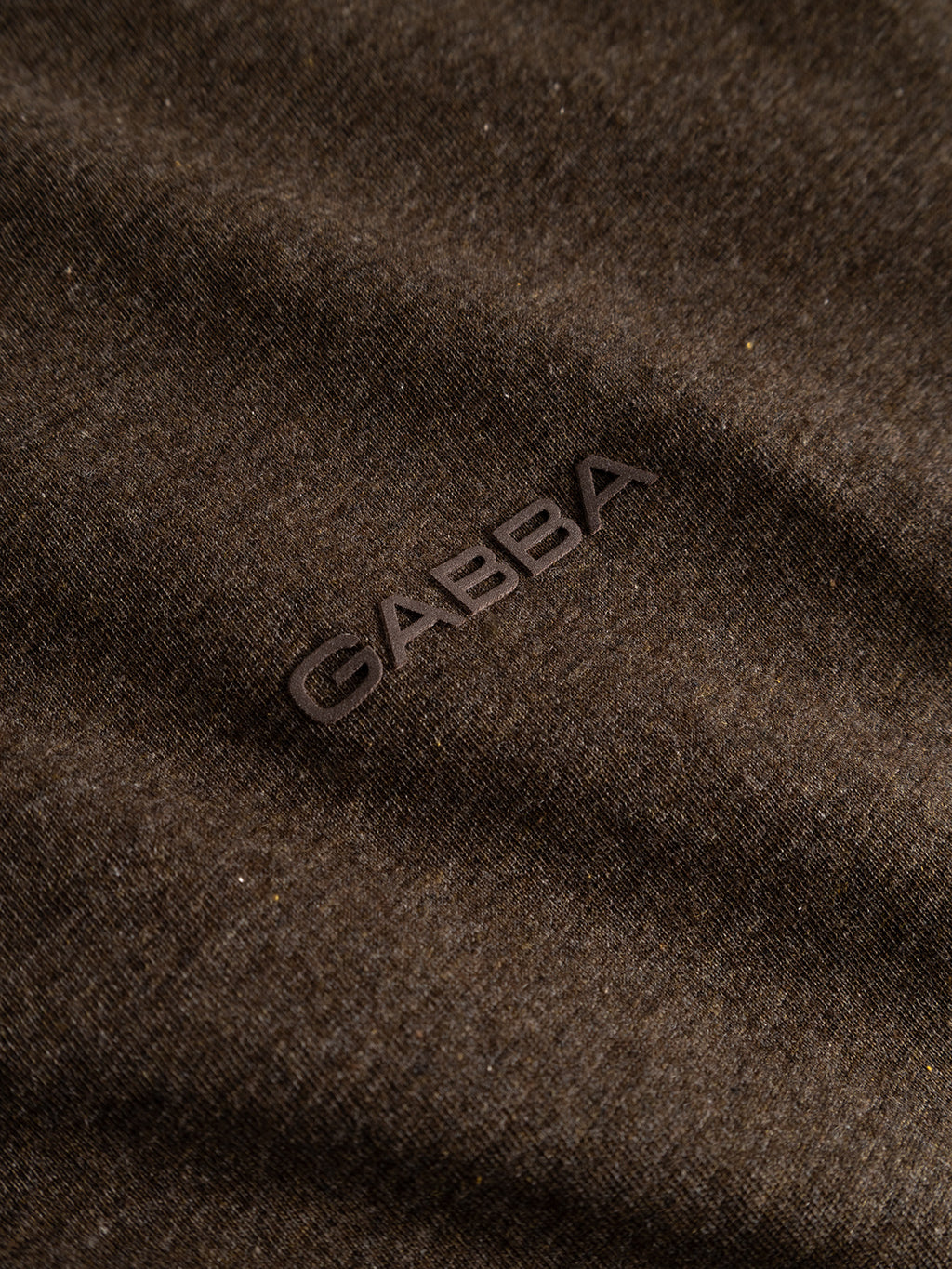 Close-up of the Gabba GABdune Melange New SS T-Shirt in Brown Slate, showcasing its soft, slightly textured melange jersey fabric and the Gabba chest logo embossed or printed in raised, dark letters on the surface. The material appears comfortable and typical of a regular fit T-shirt.