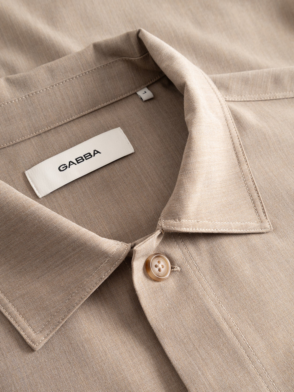A close-up of the GABtogger Tailor Dressy Overshirt in Sand shows its beige, collared design with a fastened button. Inside the collar, a white label displays the Gabba brand name in black text. The wool-blend overshirt features a smooth, subtly textured melange fabric.