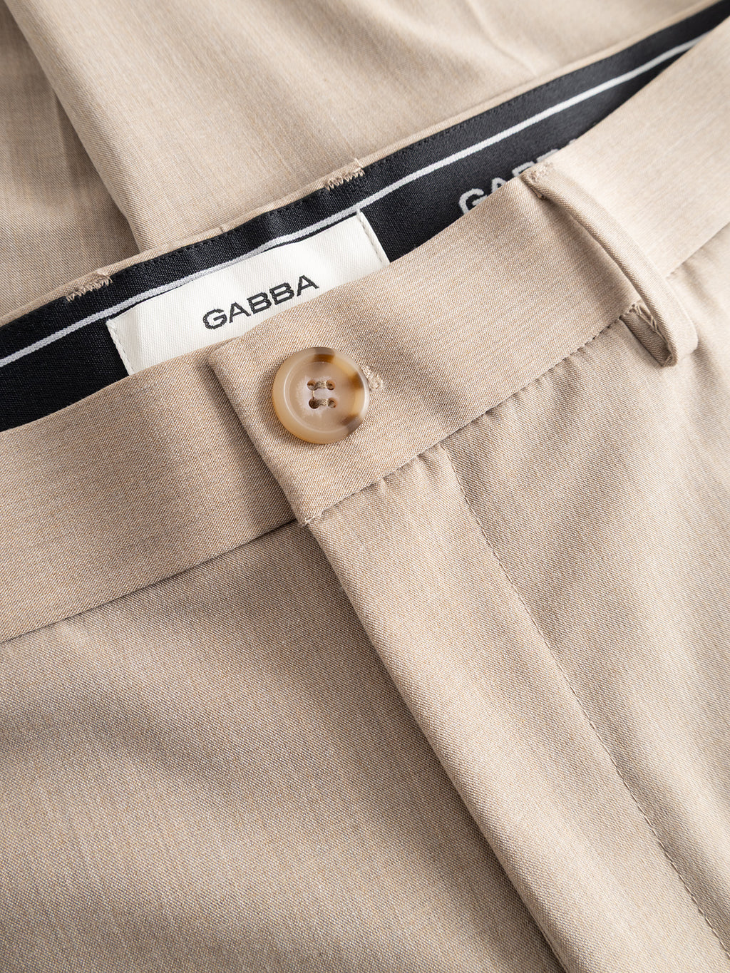 Close-up of the Gabba GABjoe Tailor Dressy Pant in sand, featuring a beige tapered fit, tan button, visible belt loop, and a black and white inner waistband with the Gabba logo. The fabric is smooth with a subtle texture.
