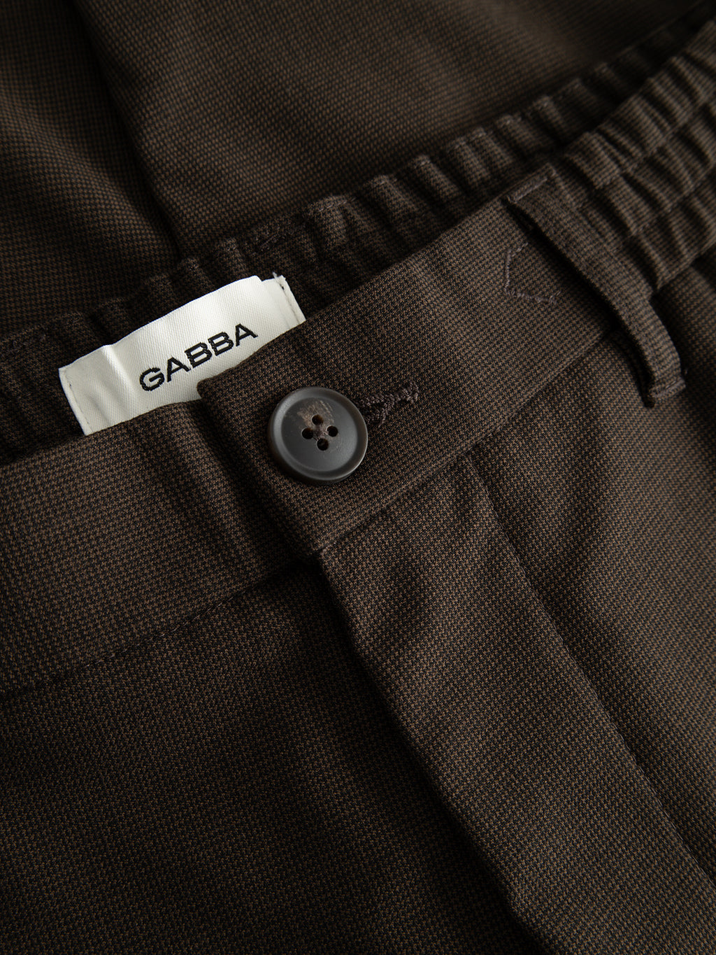 Close-up of the GABBA GABmonza Tasca Deme Pant in Brown Slate, featuring a button and an elastic waistband, with a white Gabba label. The relaxed straight fit showcases the subtle micro-patterned fabric for added texture.