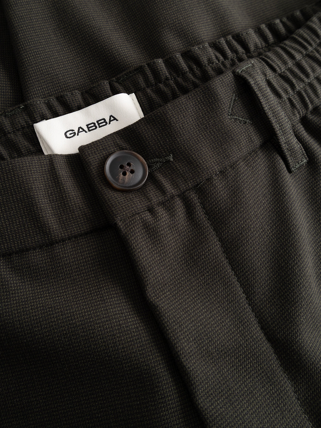 Close-up of the GABmonza Tasca Deme Pant in Forest Night by Gabba, featuring dark-colored fabric with a relaxed straight fit. The pants showcase an elastic waistband, a black button, and a white Gabba label. The micro-patterned material adds subtle texture to the design.