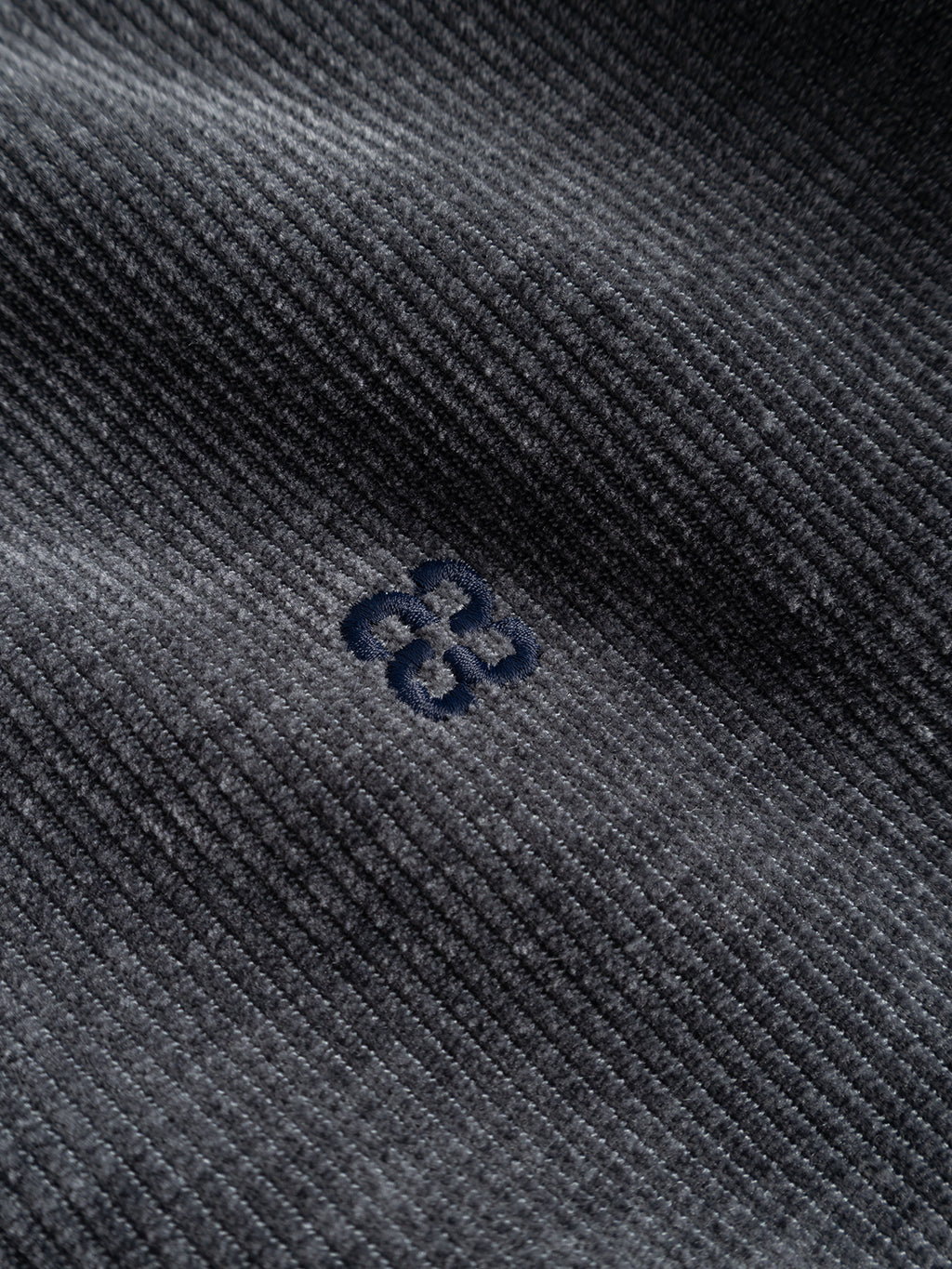 Close-up of the GABnik Pulz Fune Jacket by Gabba in Turbulence Blue, showcasing its dark gray jersey corduroy fabric with clearly visible vertical ridges. A small, subtle navy blue embroidered geometric design is featured at the center, highlighting the modern outerwear aesthetic.