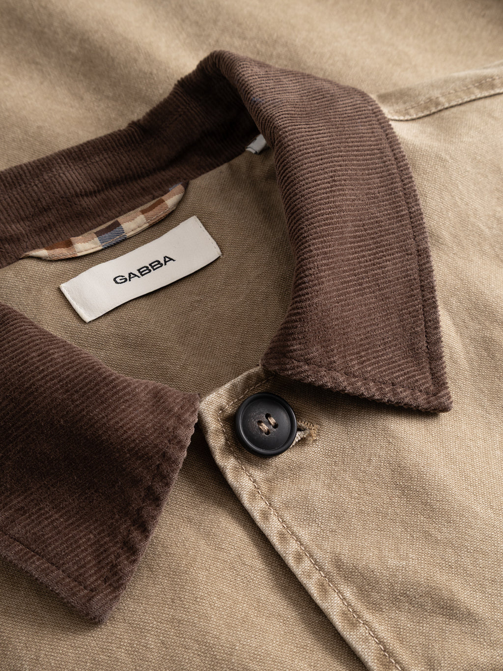 Close-up of a brown corduroy collar on the GABotto Tela Outerwear - Sand Crocker jacket by Gabba, crafted from tan washed cotton canvas. The jacket features a black button and a white label reading “GABBA,” with a plaid inner lining partially visible at the neckline.