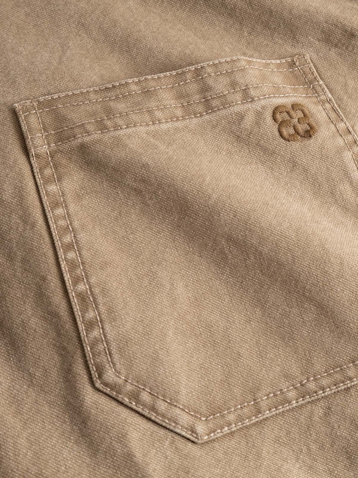 A close-up of a beige fabric pocket on the Gabba GABotto Tela Outerwear - Sand Crocker jacket, crafted from washed cotton canvas with visible stitching and a small embroidered logo featuring two overlapping C letters in the top right corner.