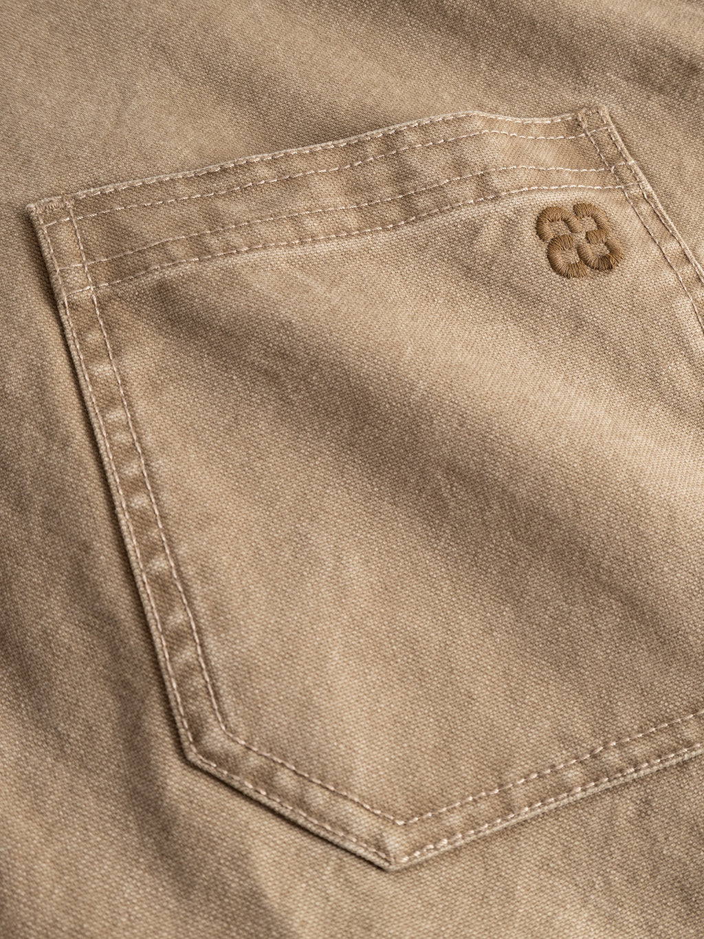 A close-up of a beige fabric pocket on the Gabba GABotto Tela Outerwear - Sand Crocker jacket, crafted from washed cotton canvas with visible stitching and a small embroidered logo featuring two overlapping C letters in the top right corner.
