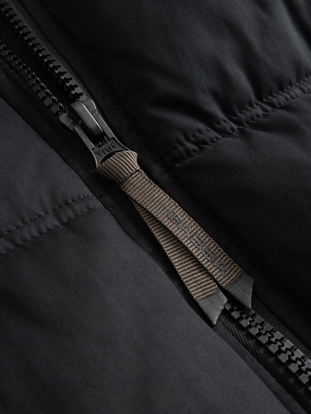 Close-up of a black GABjulien Calle Outerwear padded insulation jacket, featuring a branded fabric pull tab on the metal zipper slider.