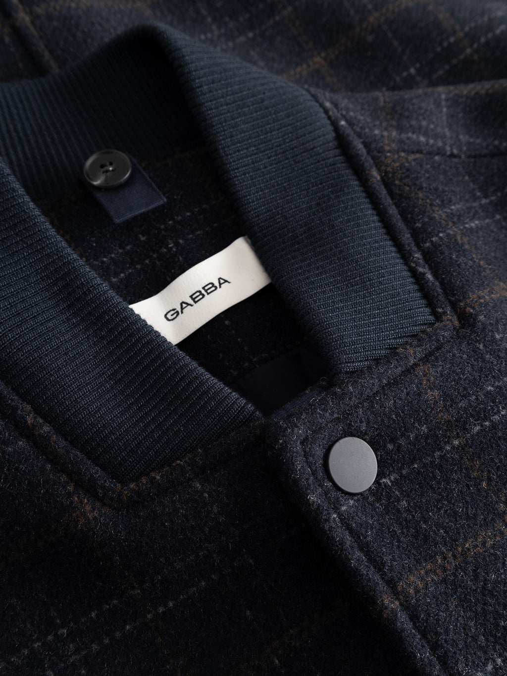 Close-up of the Gabba GABmatheo Check Bomber Jacket—a men’s outerwear piece in a blue check pattern, featuring a ribbed collar, black snap buttons, and a white Gabba label inside the collar.