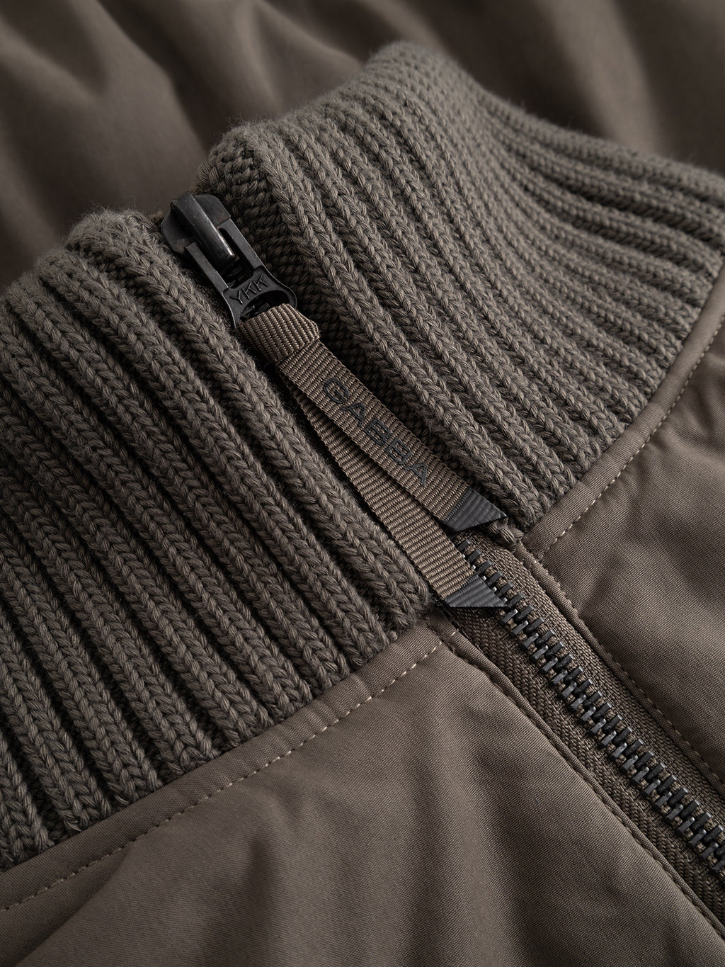 Close-up of the Gabba GAByuri Calle Outerwear in Brown Slate, showcasing a ribbed knit collar with a partially open zipper. The visible zipper pull features finely engraved text, highlighting the jacket’s attention to detail. This modern piece offers a boxy, relaxed fit that blends comfort with contemporary style for an elevated wardrobe staple.