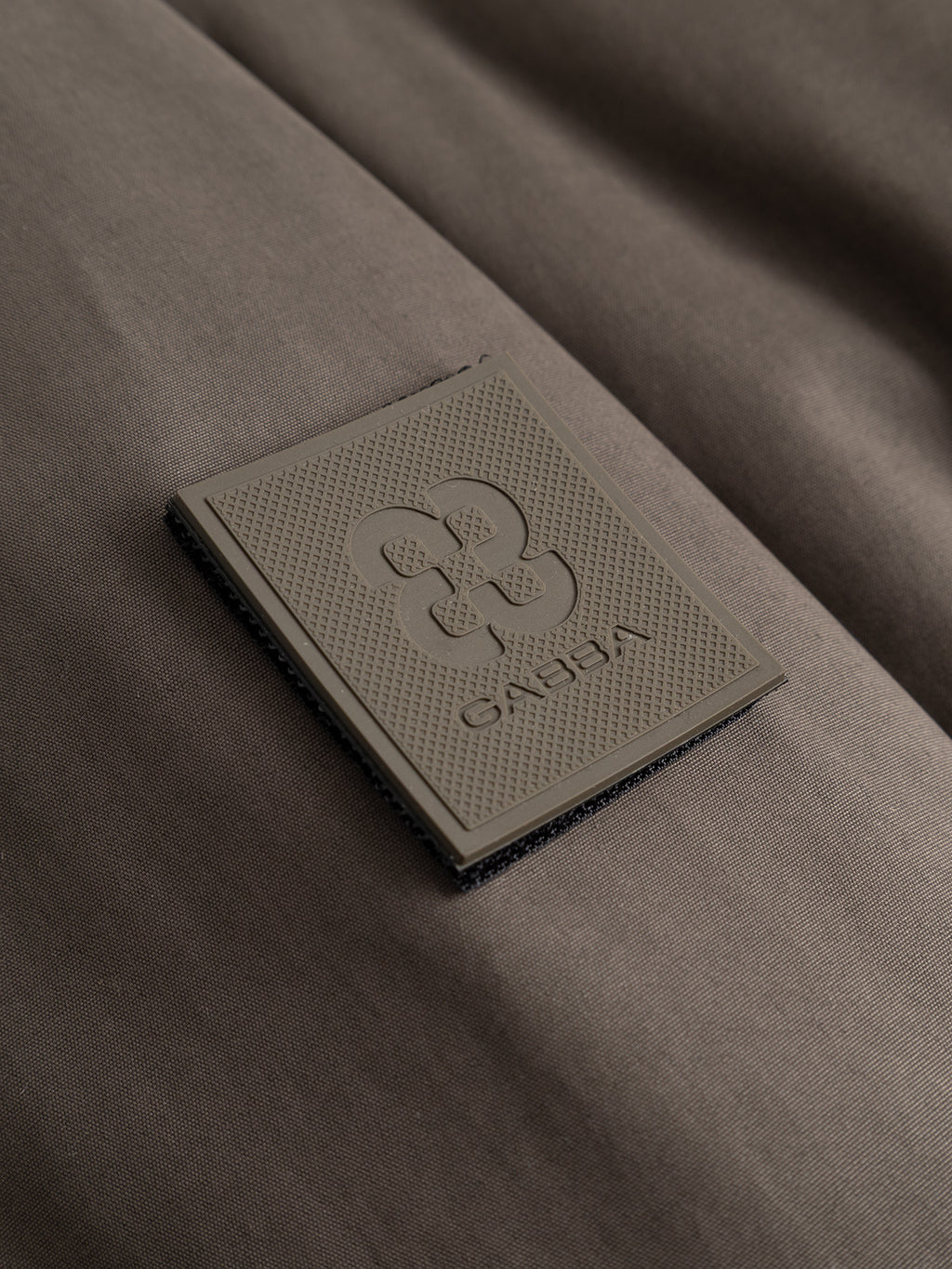 A close-up of a textured gray patch featuring a geometric logo and the word Gabba on brown fabric. The patch appears to be sewn onto the material, adding a distinctive detail to the GAByuri Calle Outerwear - Brown Slate by Gabba.