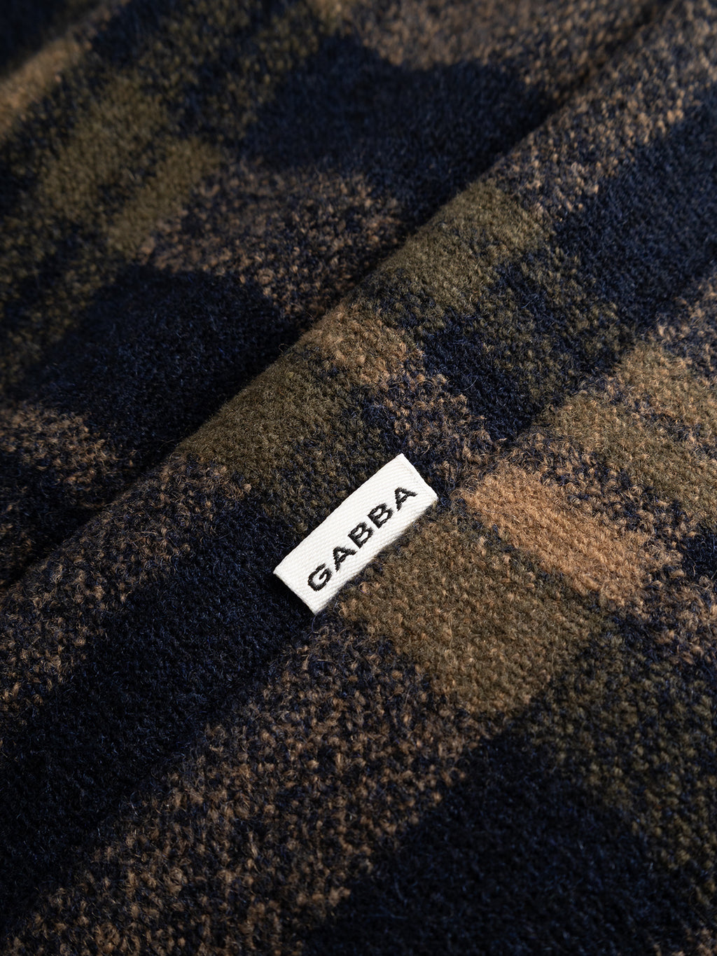A closeup of the soft, textured fabric of the Gabba GABleeds Chezy Coat in Forest Night reveals refined quality in dark and light brown tones, accented by a small white label reading “Gabba”—an outerwear essential with a timeless, tailored silhouette.