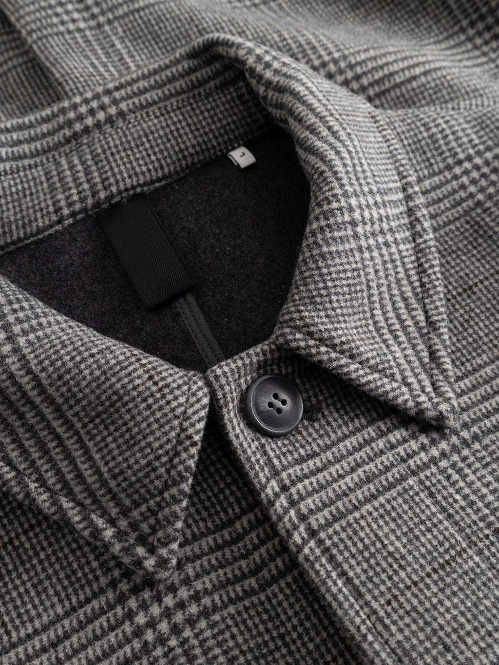 Close-up of the Gabba GABowen Scacco Coat in Grey Check, highlighting its pointed collar, black button, and visible size tag. Expertly crafted from premium wool-blend checked fabric, this tailored coat features a classic grey checkered pattern with fine lines and a soft, refined texture.